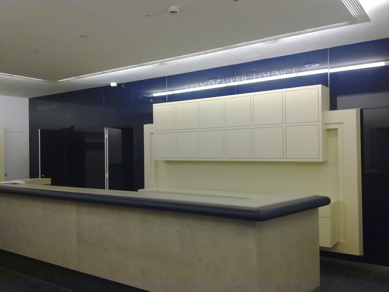 Moderne Empfangstheke in einem Foyer: heller Tresen mit dunkler Kante, dahinter eine cremefarbene Schrankwand vor dunkelblauen Paneelen und beleuchteter Decke; links ein Eingang.